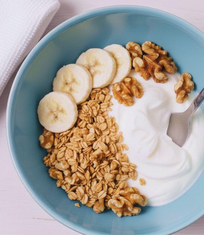 Una ciotola di yogurt con granola, banane a fette e noci, appoggiata su un tavolo di legno rosato chiaro. Accanto alla ciotola c'è un asciugamano bianco piegato. Un cucchiaio di metallo è immerso nello yogurt.