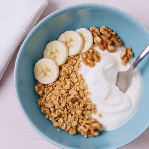 Una ciotola di yogurt con granola, banane a fette e noci, appoggiata su un tavolo di legno rosato chiaro. Accanto alla ciotola c'è un asciugamano bianco piegato. Un cucchiaio di metallo è immerso nello yogurt.